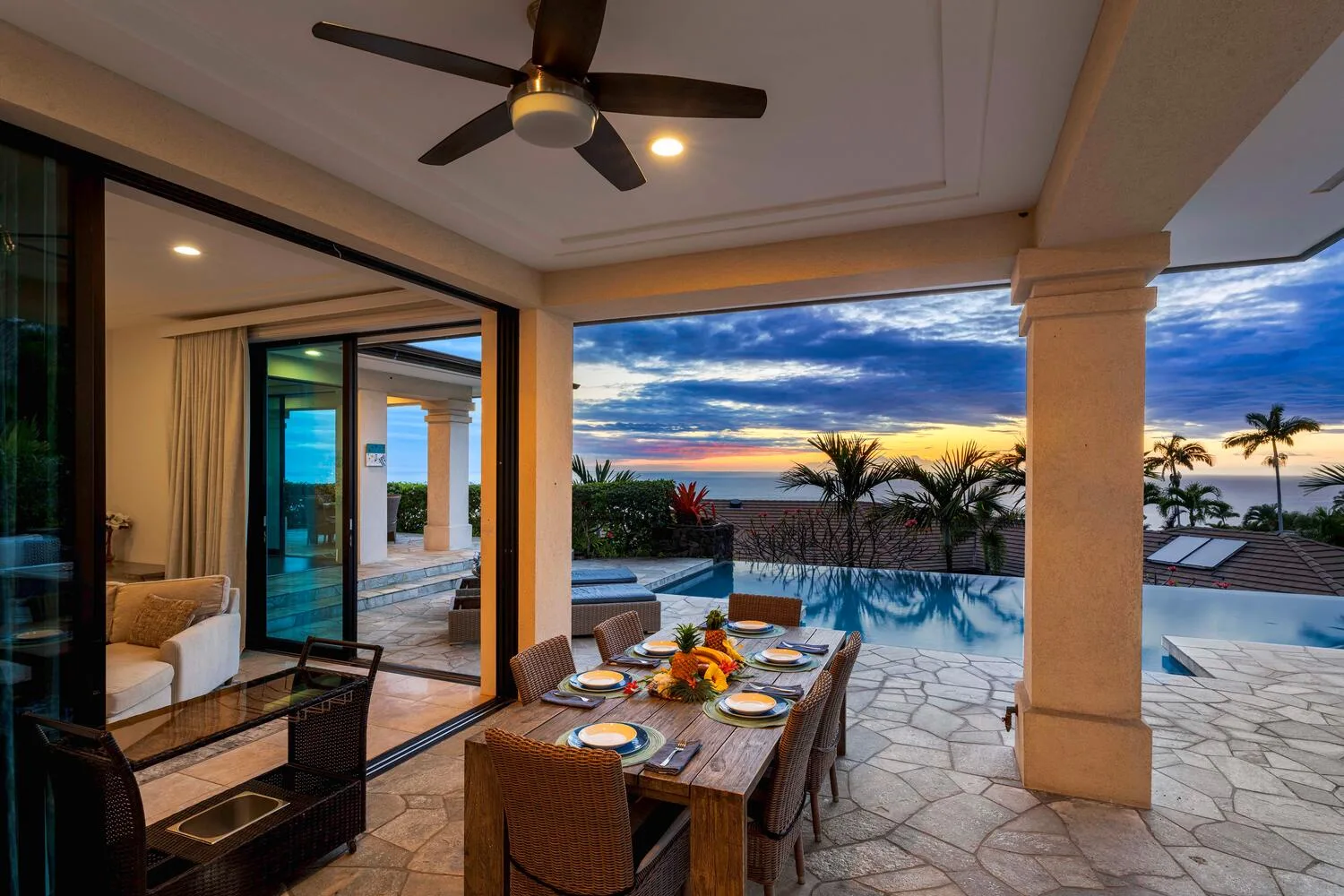 A covered patio dining area opens onto a pool and a colorful sunset view.