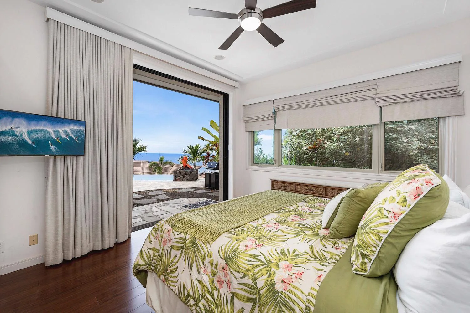 A bedroom with a floral bedspread, a ceiling fan, and windows overlooking greenery.