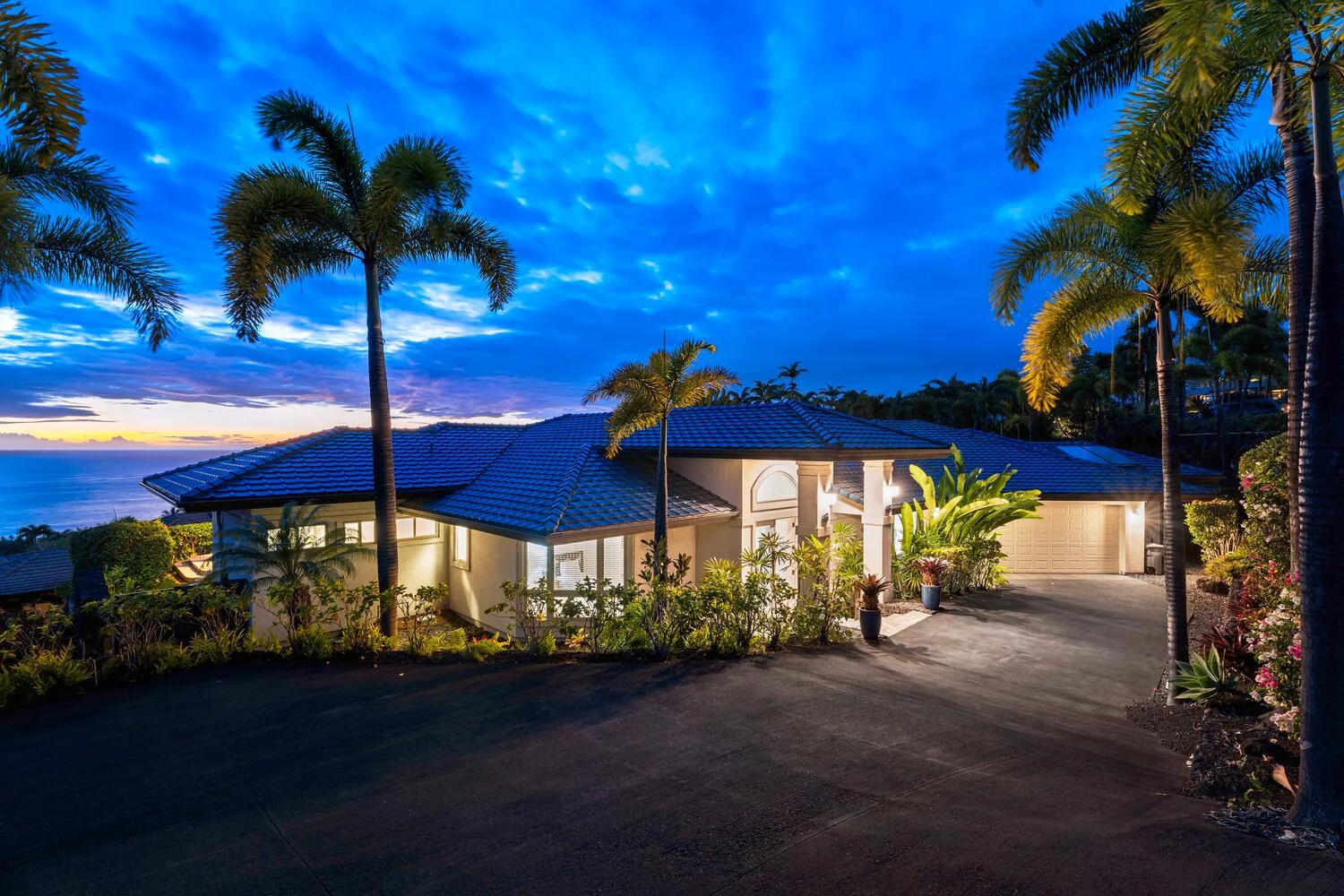 A single-story house with a tiled roof sits beside a palm-lined driveway at dusk.