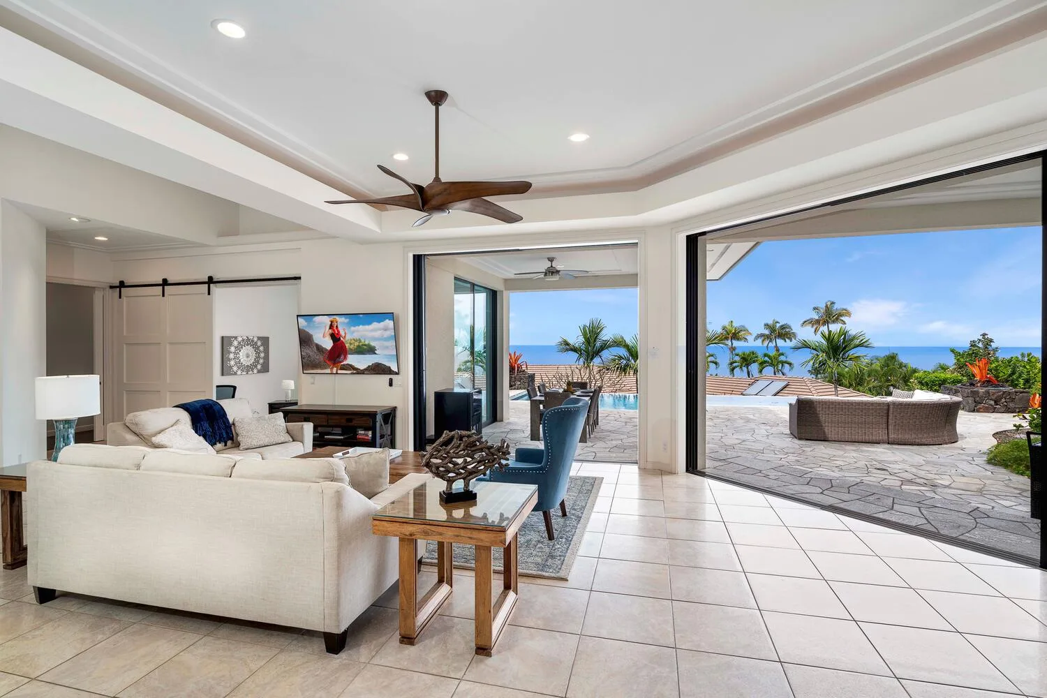 A bright living room opens onto a patio with palm trees and an ocean view.