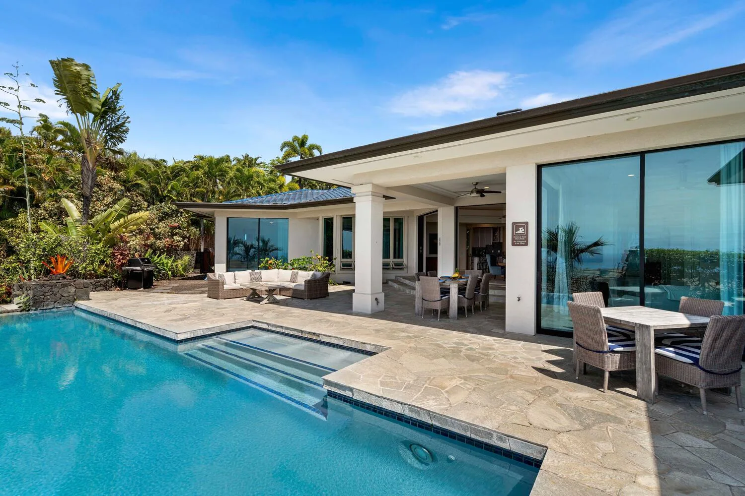 A modern patio beside a swimming pool, with outdoor seating and tropical greenery.