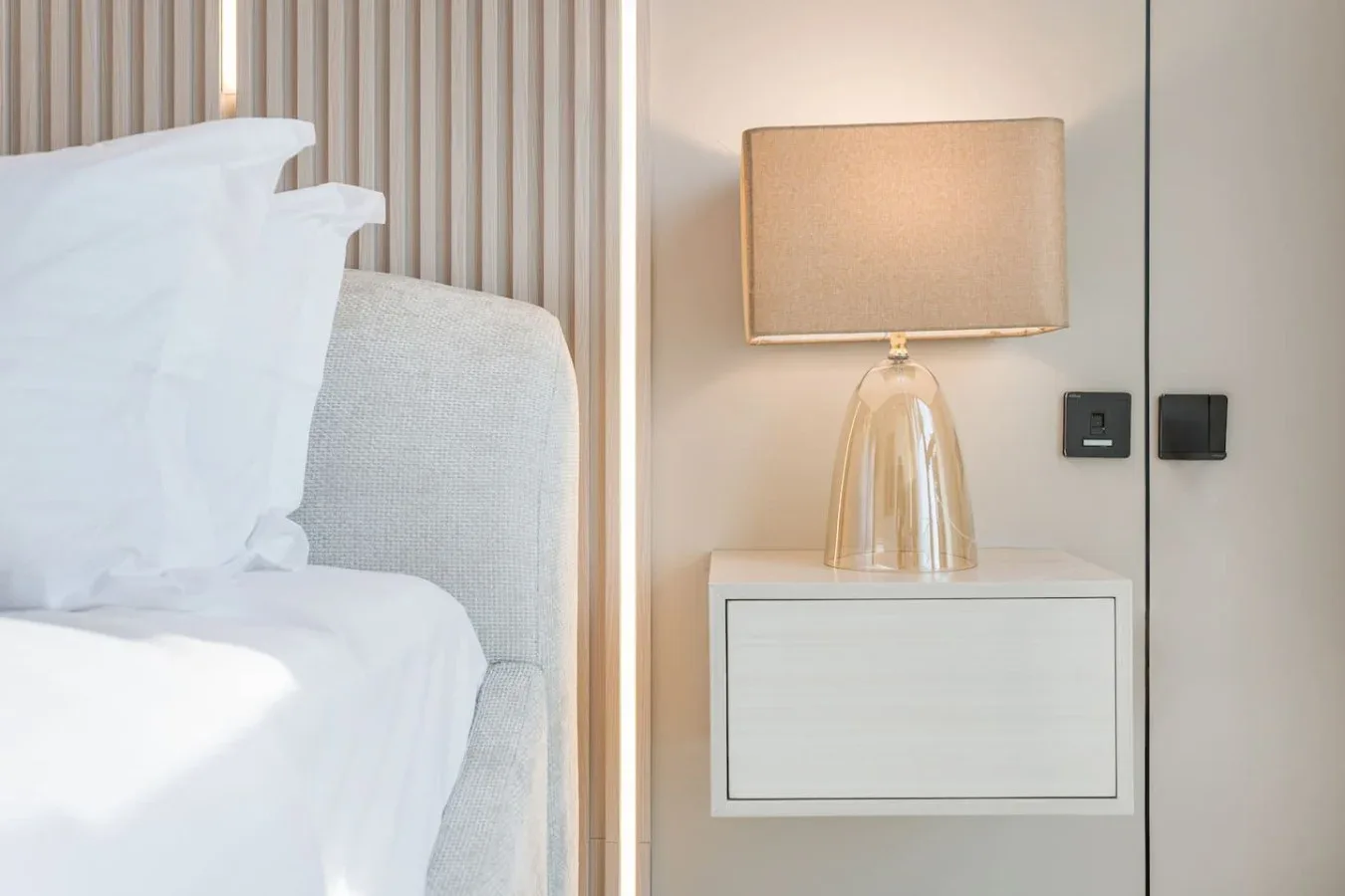Un dormitorio con ropa de cama blanca junto a una mesita y una lámpara.