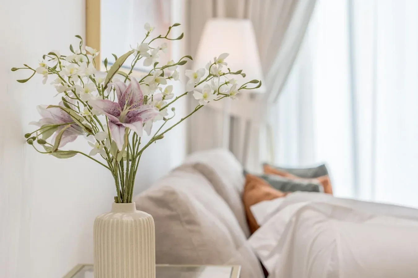 Un dormitorio con luz suave, una cama con ropa blanca y flores sobre una mesa auxiliar.
