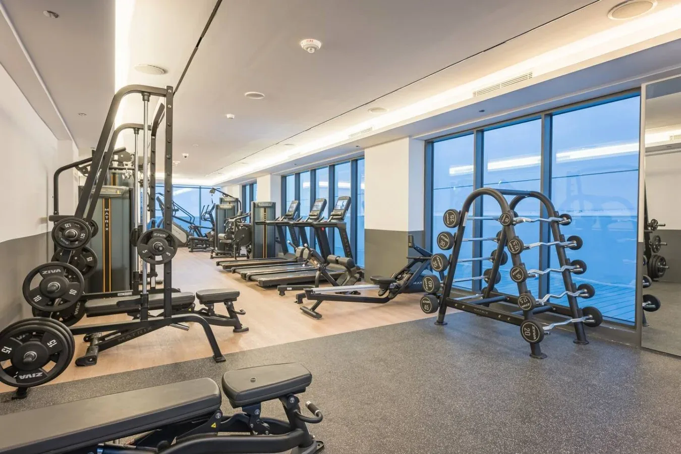 Un gimnasio con máquinas de ejercicio y soportes de pesas junto a grandes ventanales.