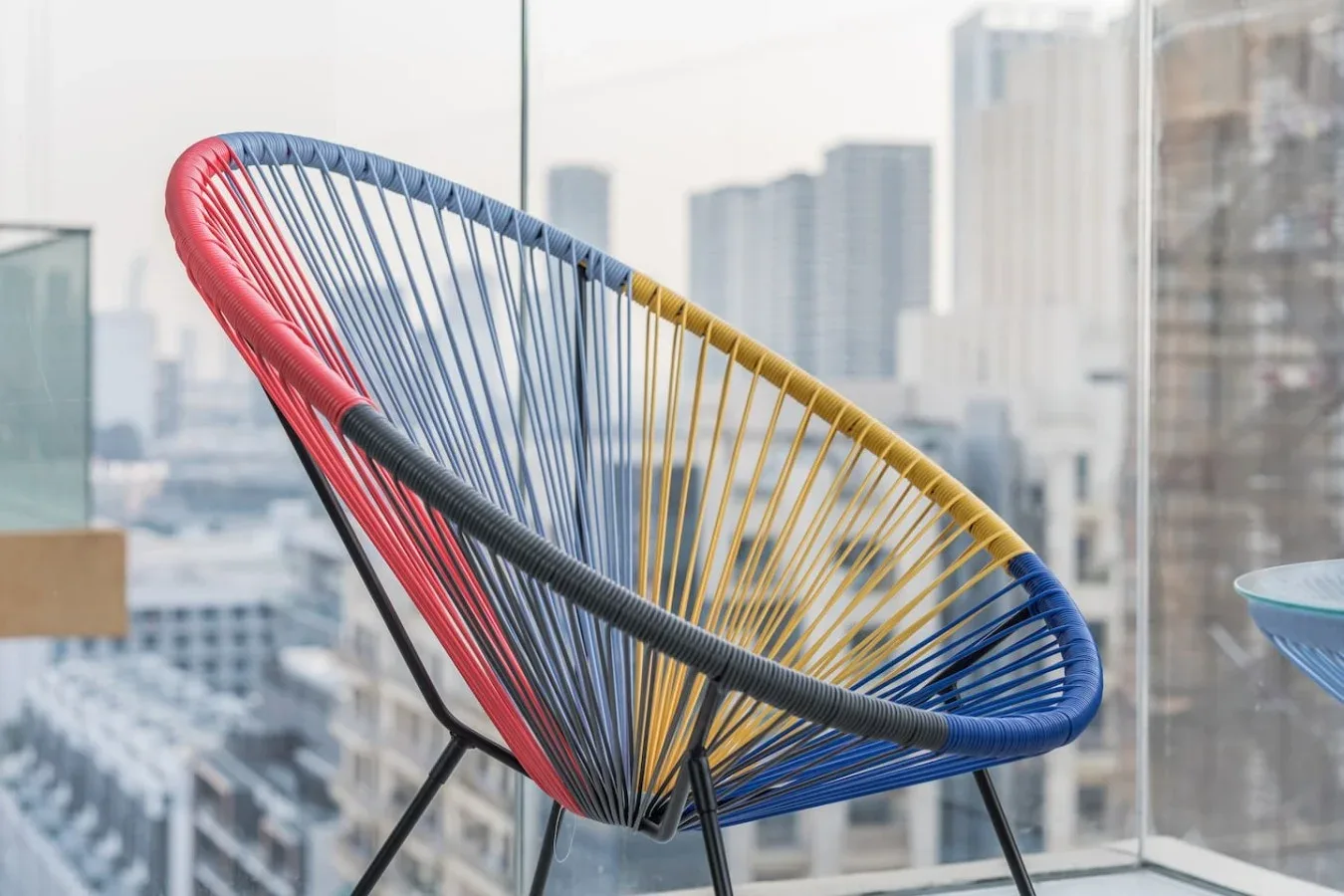 Una silla tejida de colores está junto a una ventana amplia con vistas a edificios de la ciudad.
