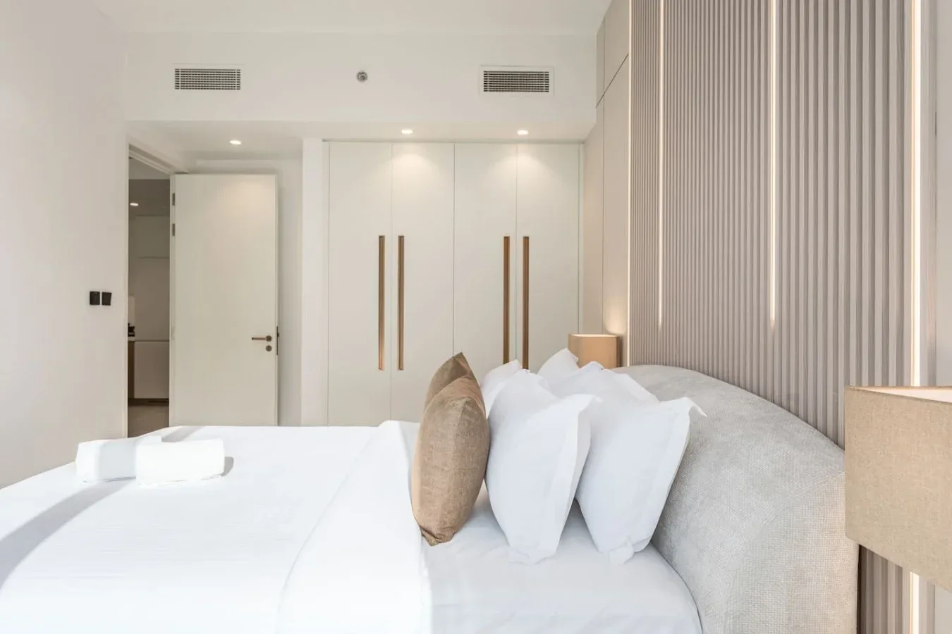 Un dormitorio luminoso con ropa de cama blanca, almohadas y un panel alto de pared estriado.