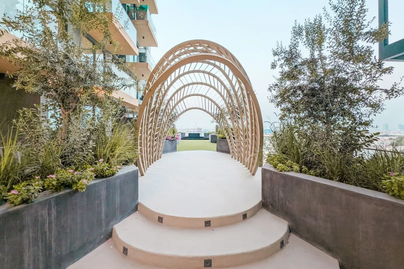 Un paseo de terraza al aire libre pasa bajo un arco curvo de madera junto a jardineras.
