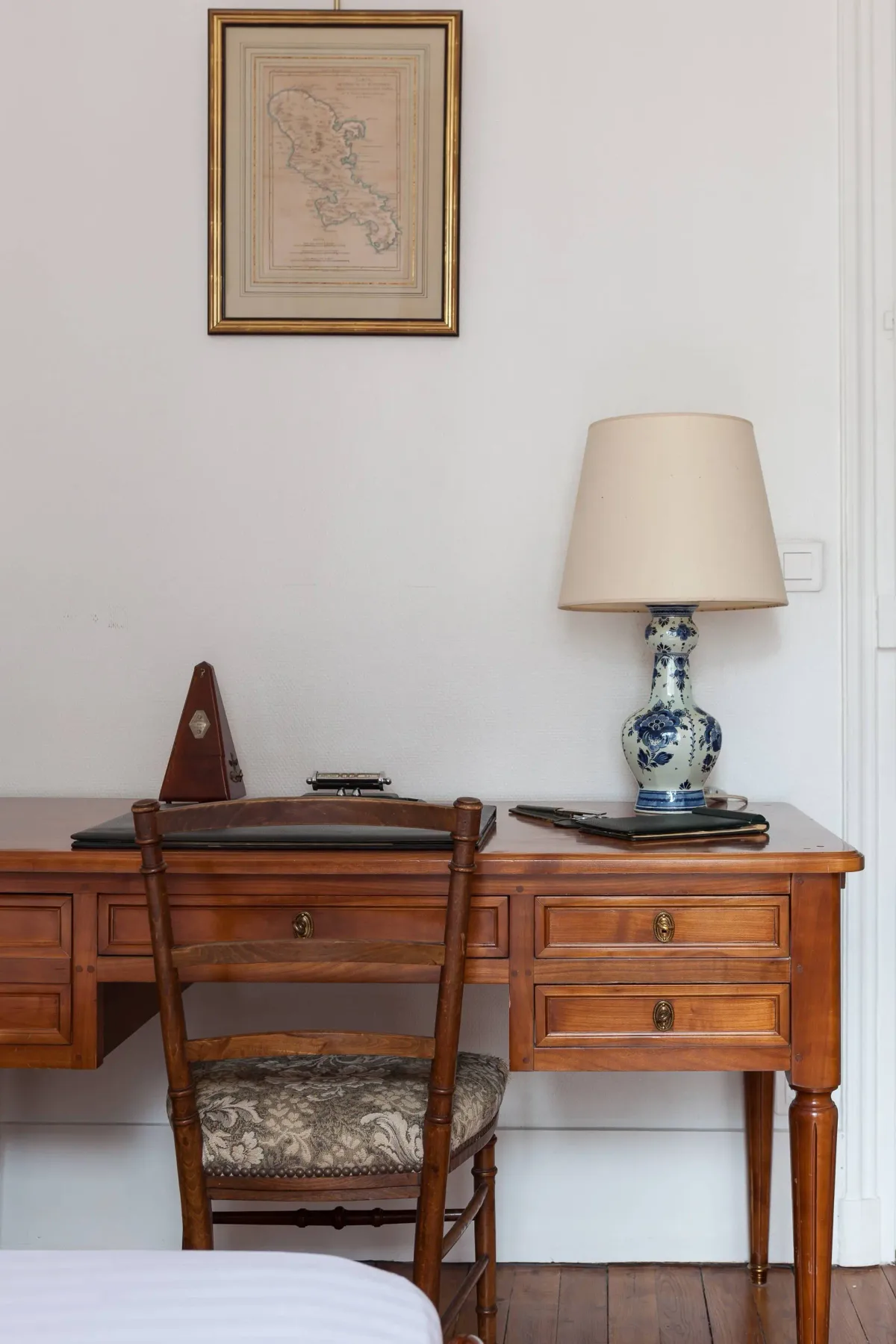 A wooden desk and chair sit beneath a framed map, with a table lamp on the desk.