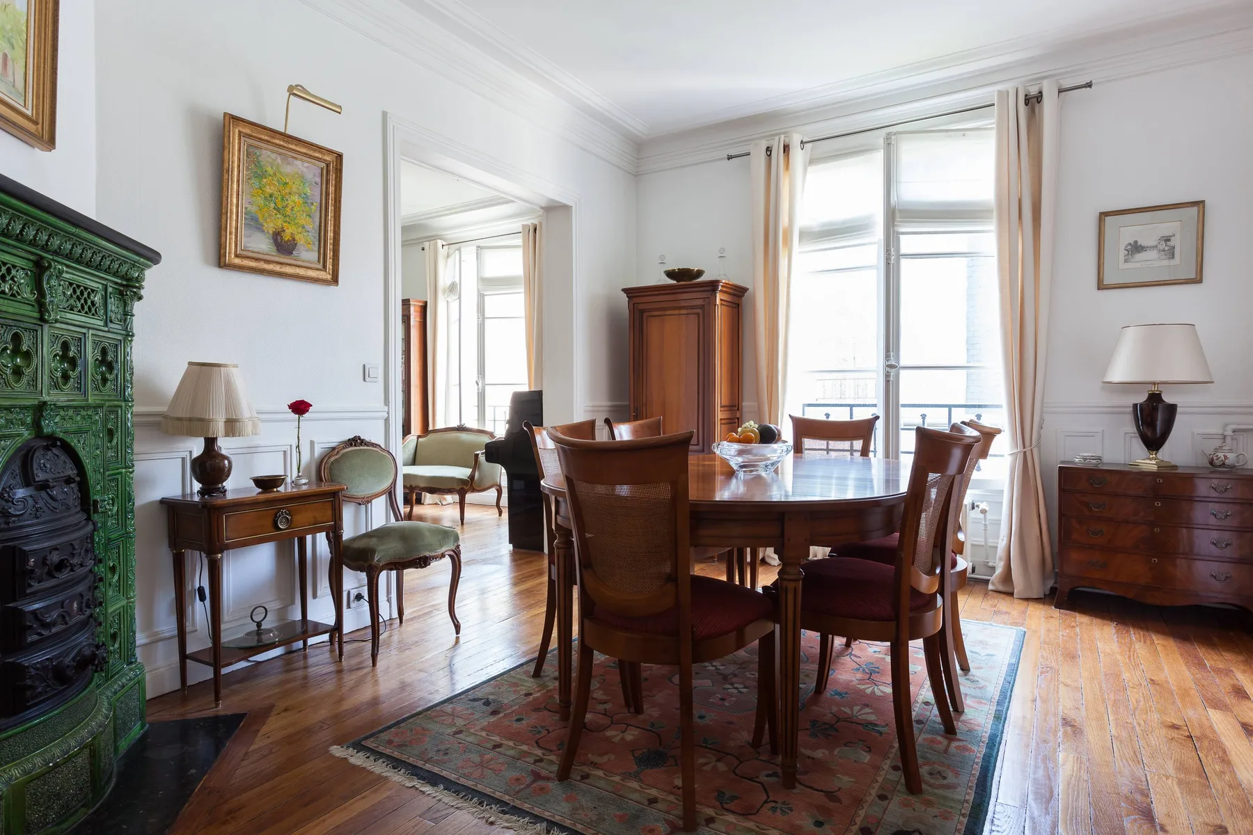 A bright dining room with a wooden table and chairs beside tall curtained windows.