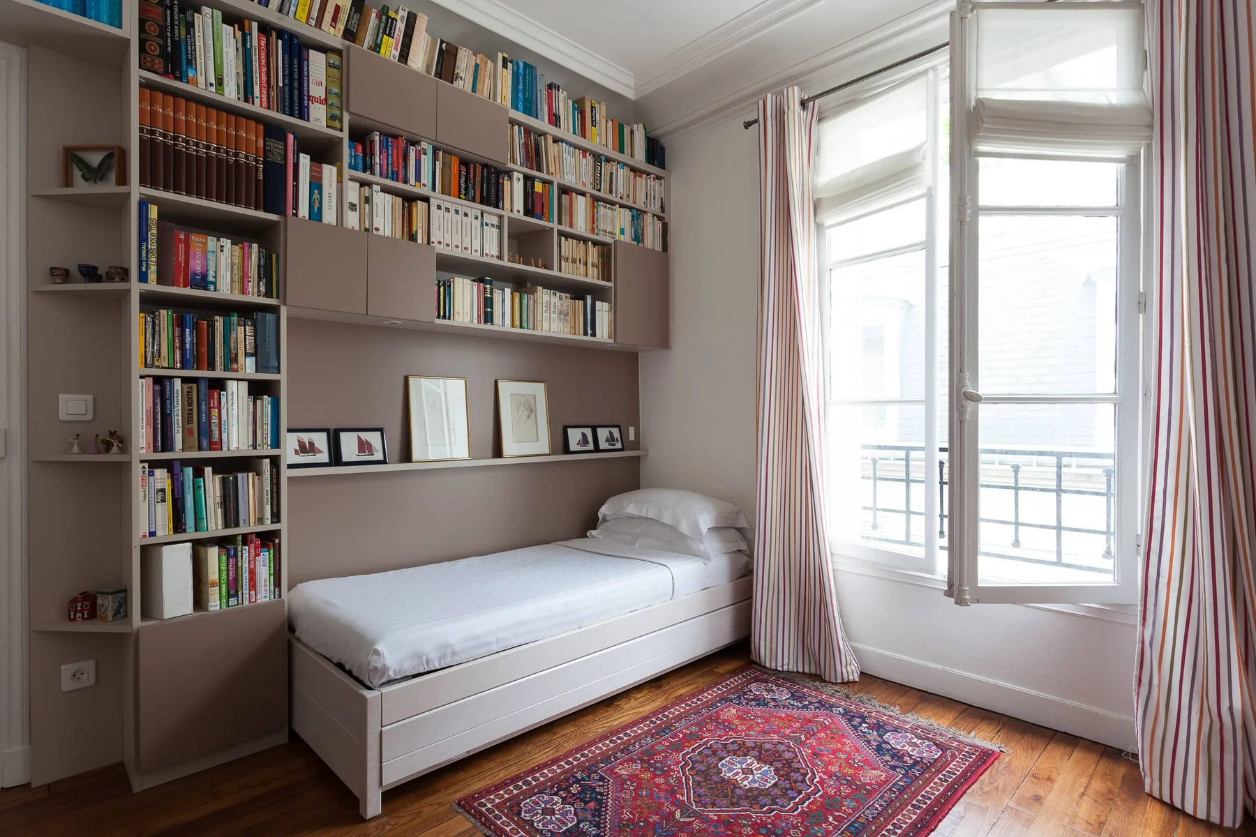 A bright bedroom with a single bed, wall-length bookshelves, and tall windows.