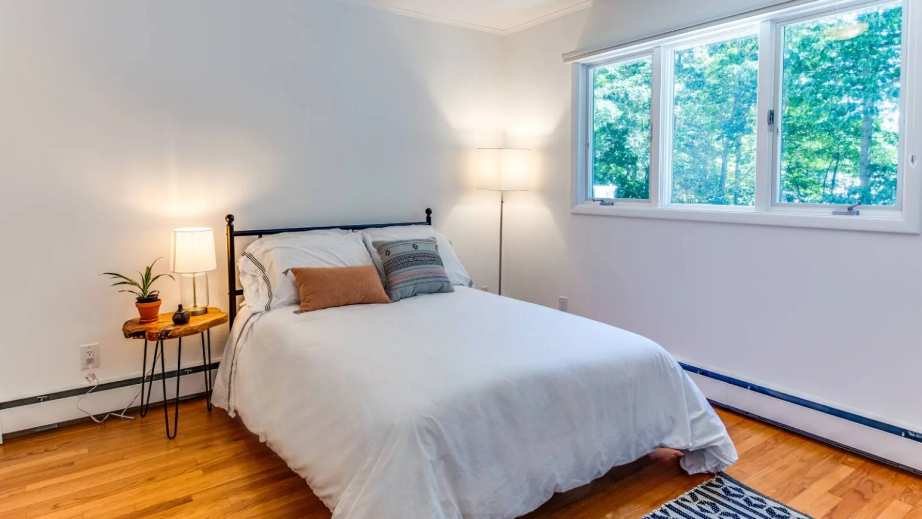 A bedroom with a bed beside a large window overlooking leafy trees.