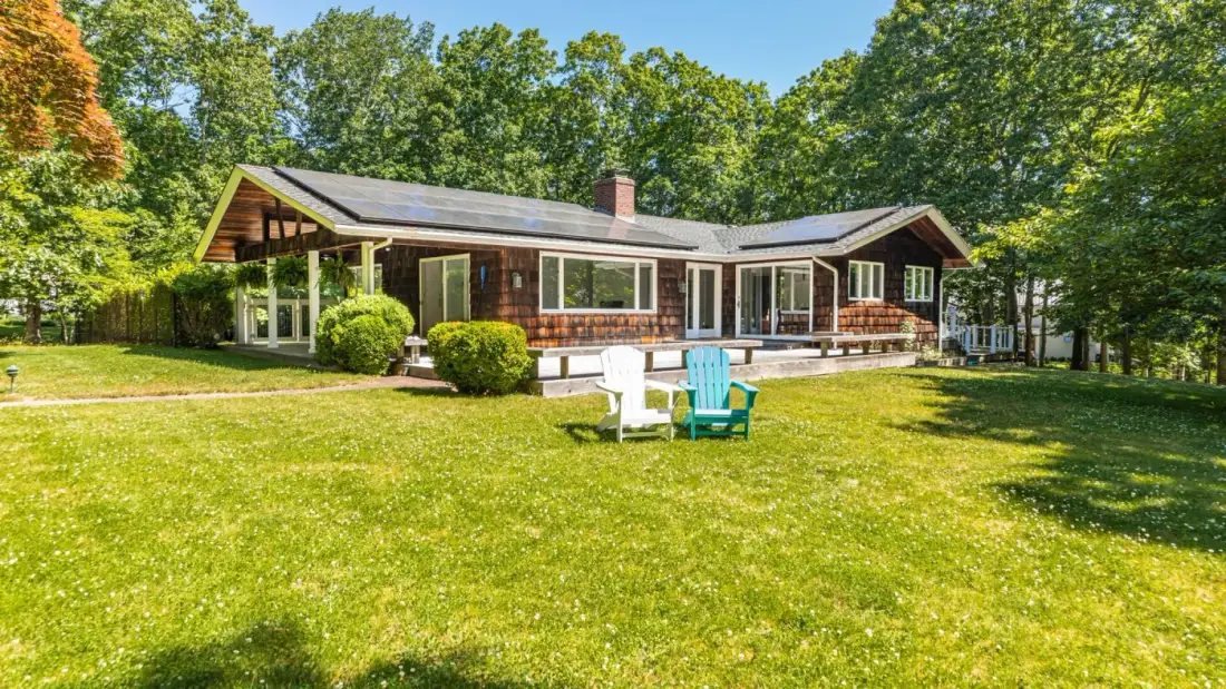 A single-story house stands beyond a broad lawn, surrounded by trees.