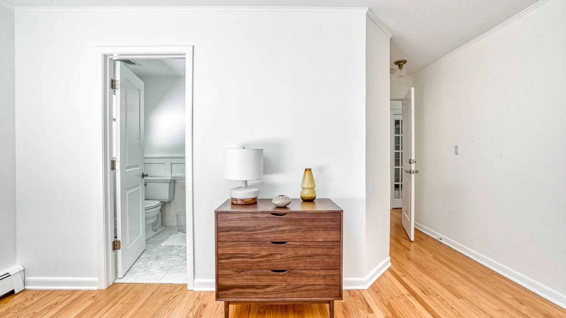 A bright bedroom area with a wooden dresser and an open bathroom doorway.