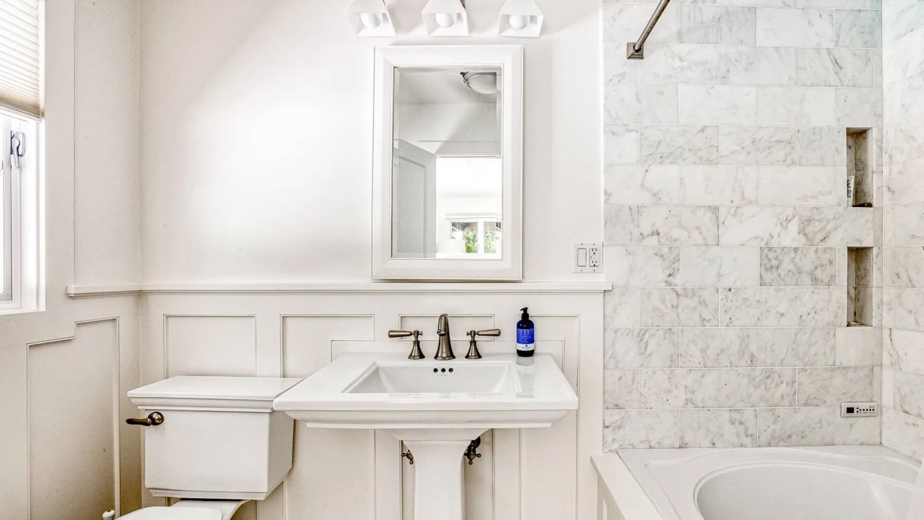 A bright bathroom with a vanity, toilet, and bathtub beside a tiled shower wall.