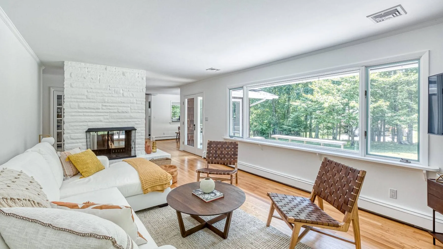 A bright living room with seating, a coffee table, and large windows overlooking trees.