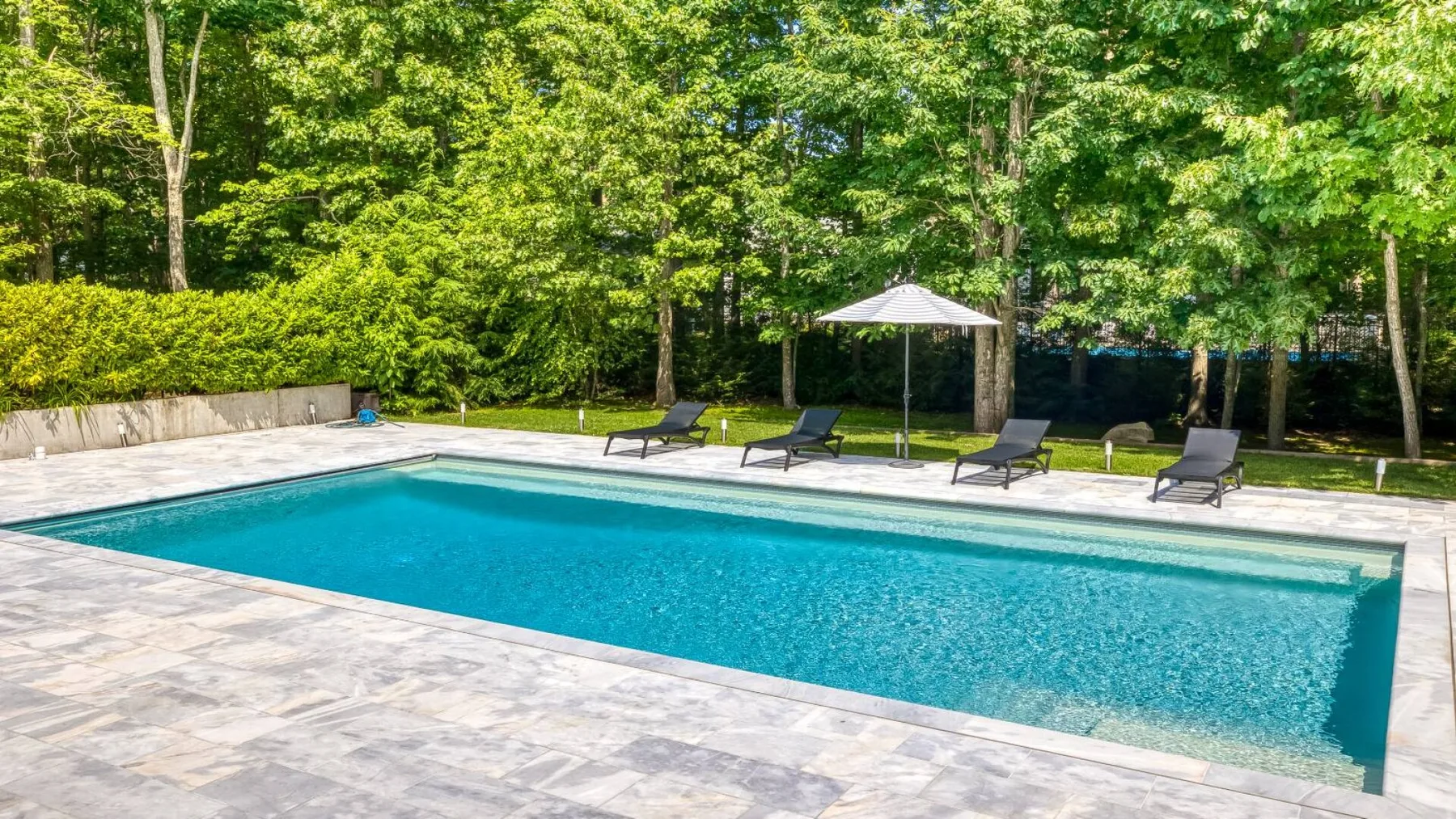 A swimming pool is surrounded by a paved deck, lounge chairs, and trees.