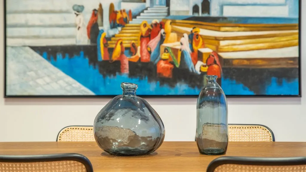 A dining table with two glass vases sits beneath a colorful painting.