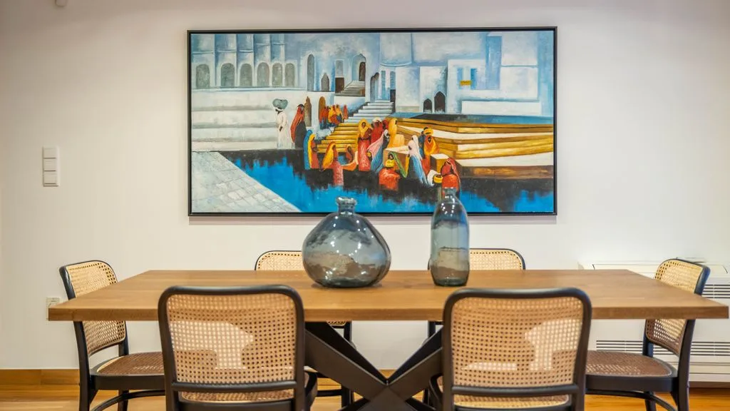 A dining area with a wooden table and a colorful framed painting on the wall.
