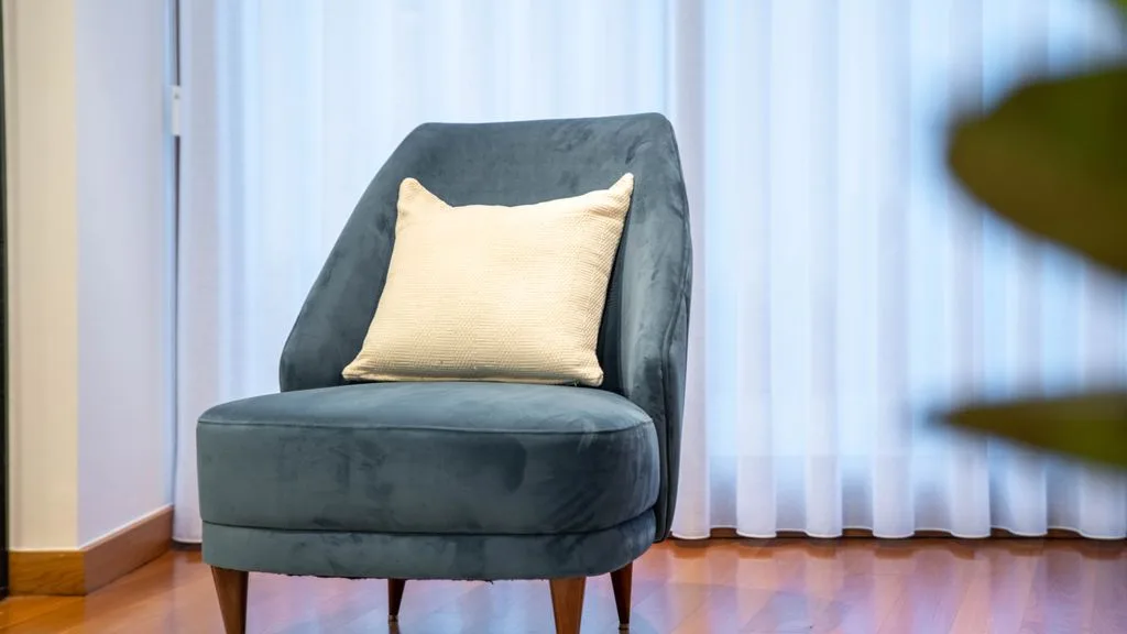 A dark upholstered armchair with a light cushion sits in front of sheer curtains.