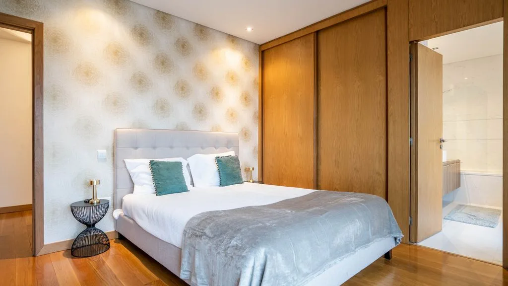 A bedroom with a double bed, patterned wallpaper, and wooden wardrobes.