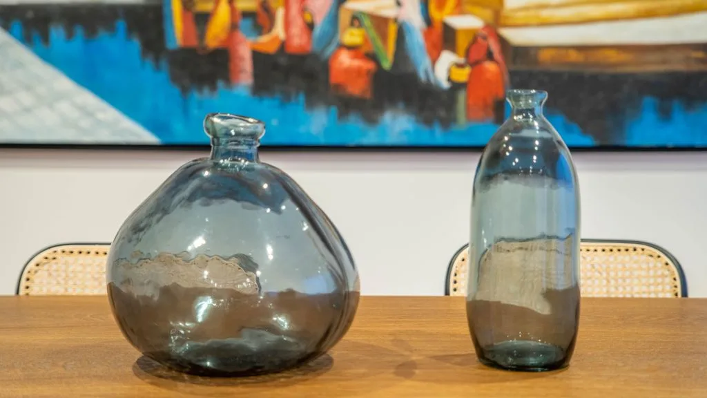 Glass vessels sit on a wooden table beneath a colorful painting.
