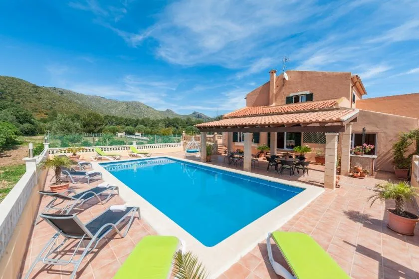 Outdoor pool beside a covered patio with seating, surrounded by hills.