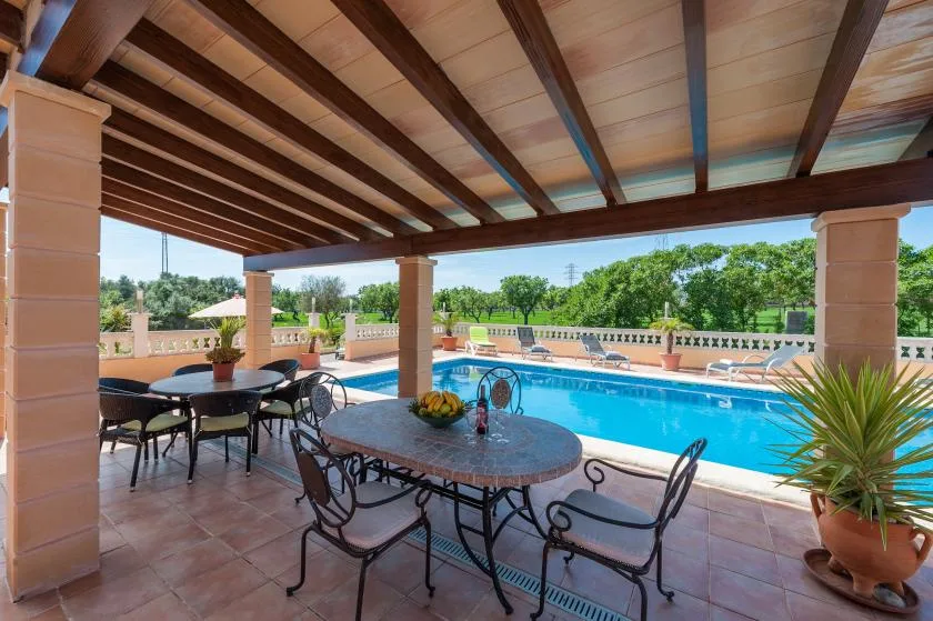 A covered patio with dining tables overlooks a swimming pool and landscaped grounds.