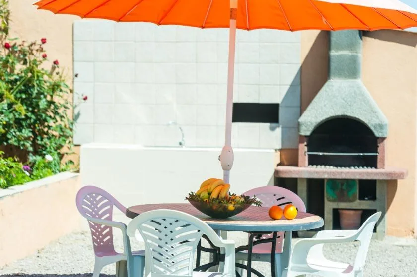 An outdoor patio dining area sits beneath an orange umbrella beside a barbecue.