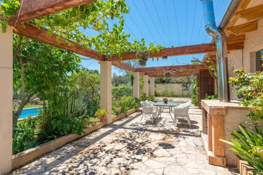 A shaded outdoor dining patio sits beneath a wooden pergola surrounded by greenery.