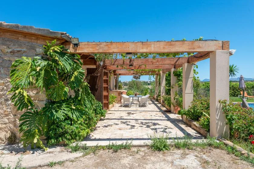 A stone patio beneath a wooden pergola leads to outdoor seating surrounded by greenery.