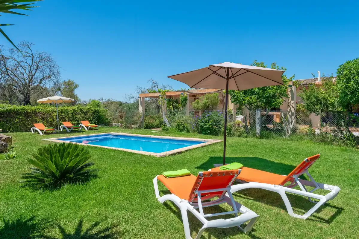 A garden pool is surrounded by green lawn, loungers, and a sun umbrella.