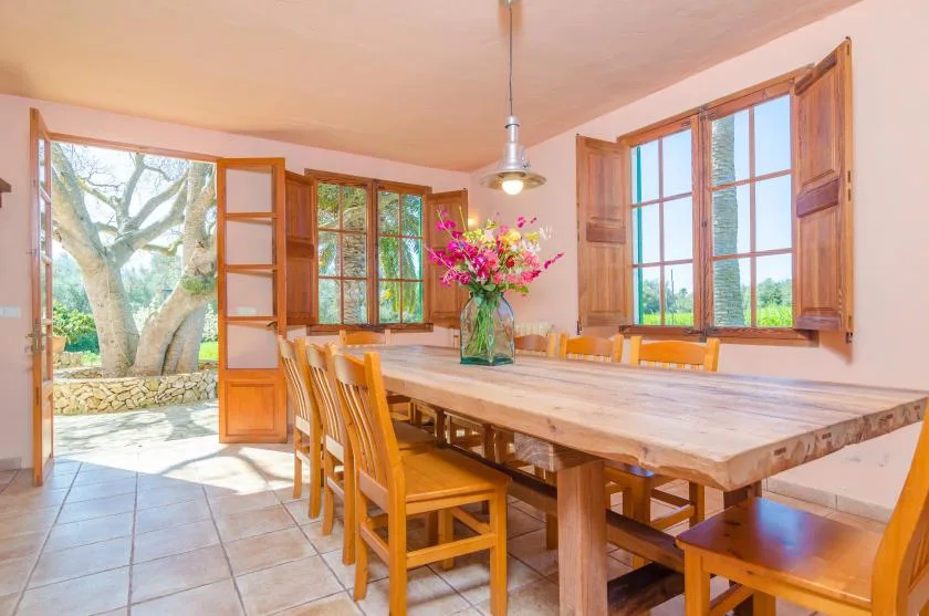 A dining area with a long wooden table and open windows overlooking greenery.