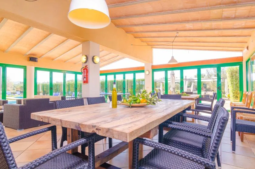 A dining area with a long wooden table and dark woven seating beside green-framed windows.