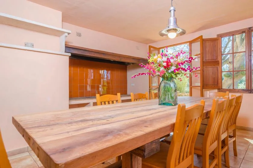 A dining area with a long wooden table, chairs, and flowers in a vase.