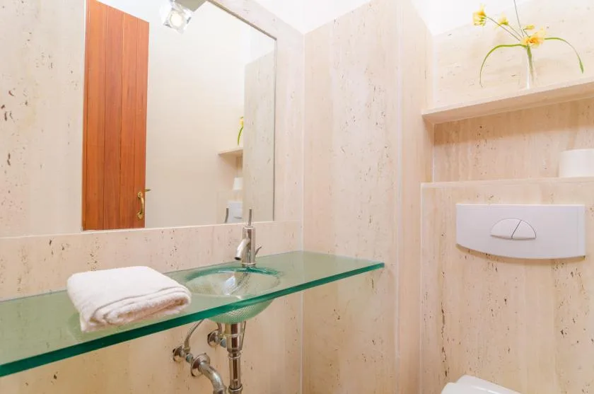 A bathroom vanity with a glass shelf, mirror, and wall-mounted toilet flush panel.