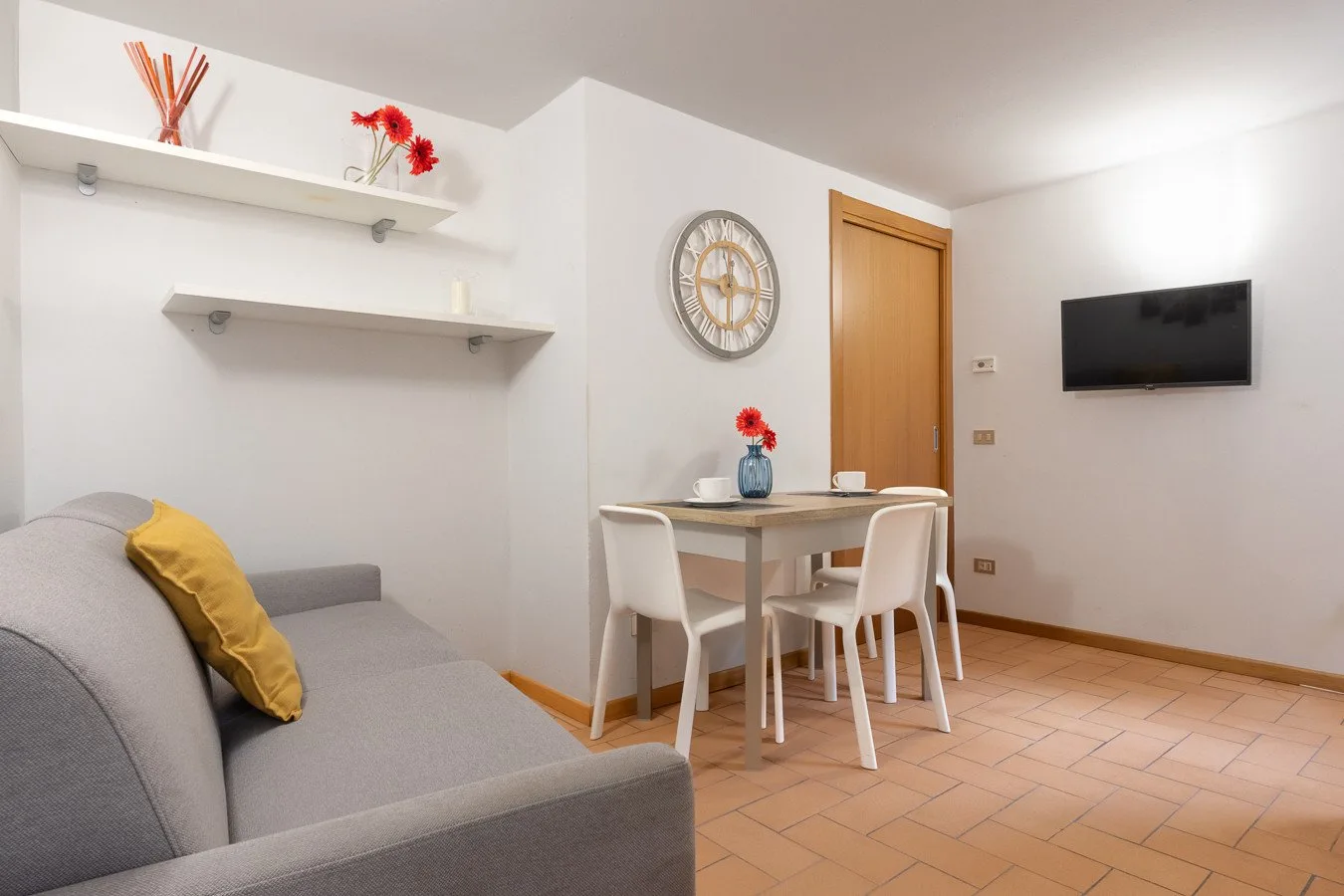 Una sala de estar con asientos grises, mesa de comedor y televisor en la pared.