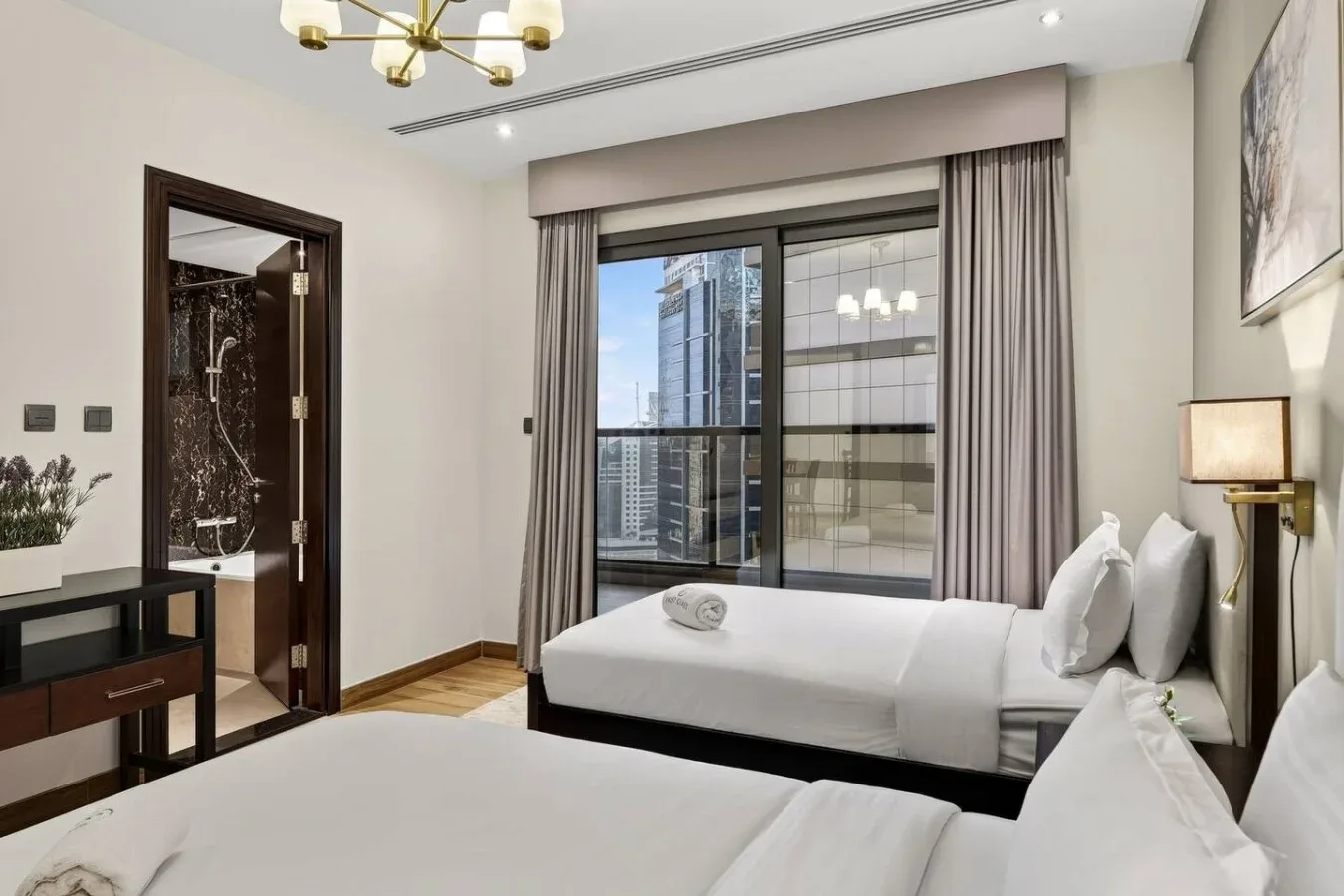Un dormitorio luminoso con ropa de cama blanca y grandes ventanas con vistas a edificios cercanos.