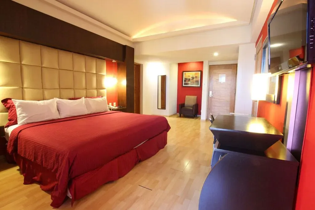Habitación de hotel con una colcha roja, dos sillones, lámparas de luz cálida y suelo de baldosas.