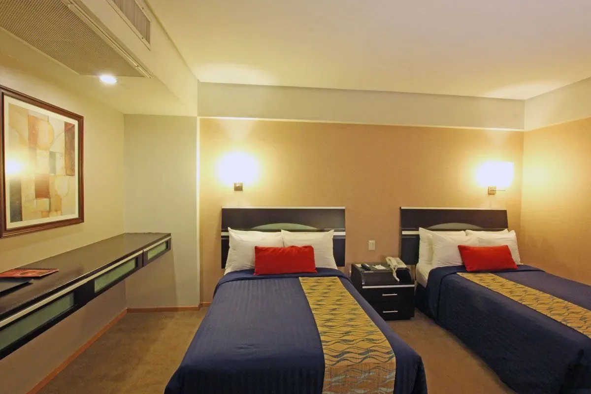 Habitación de hotel con una colcha roja, dos sillones, lámparas de luz cálida y suelo de baldosas.