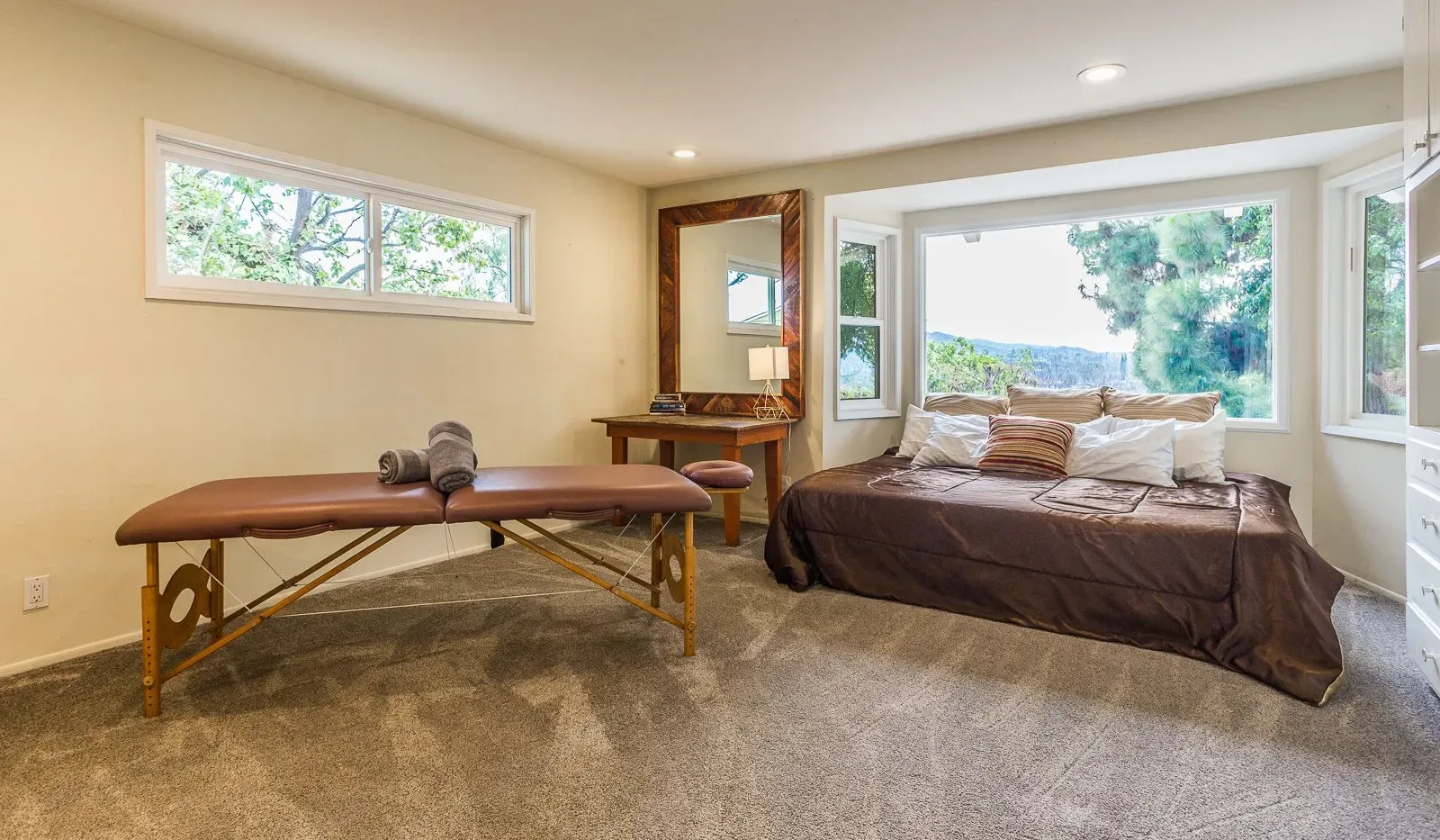 Un dormitorio con una cama y una camilla de masaje junto a ventanas grandes.