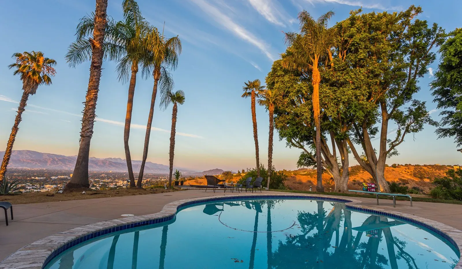 Una piscina al aire libre está rodeada de palmeras, con colinas visibles a lo lejos.