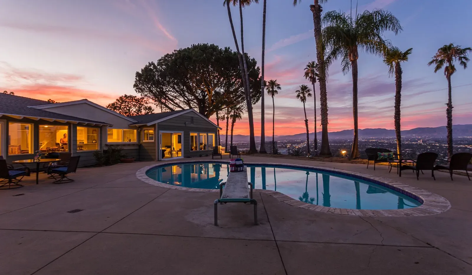 Una piscina curva está junto a una casa bajo palmeras al atardecer.