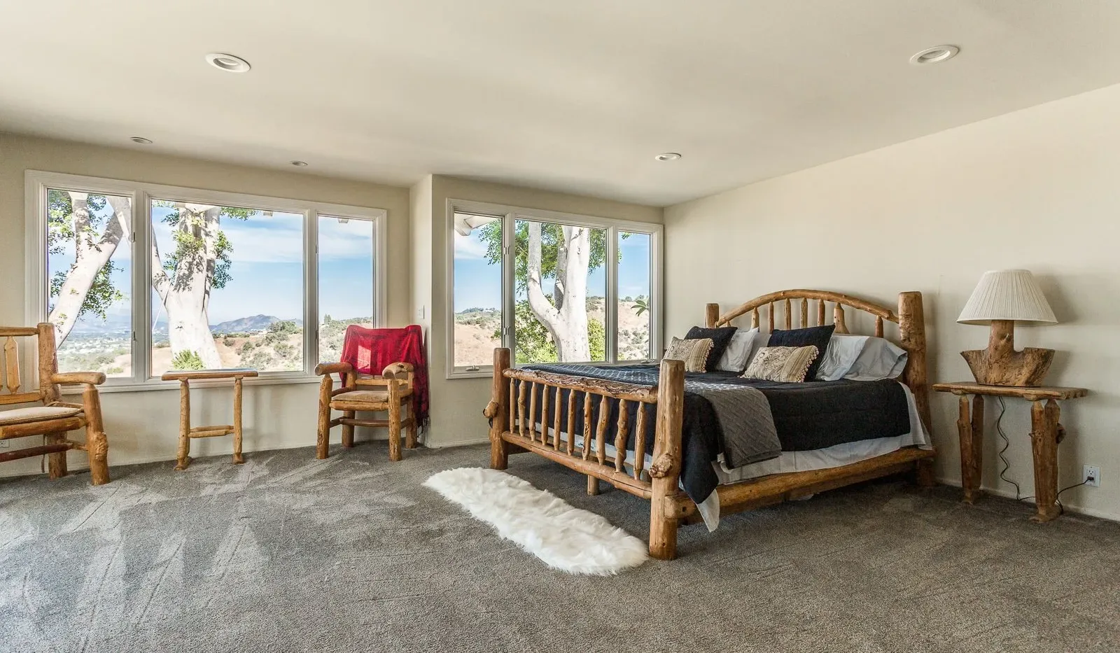 Un dormitorio con una cama de madera, asientos y grandes ventanas con vistas a árboles y agua.