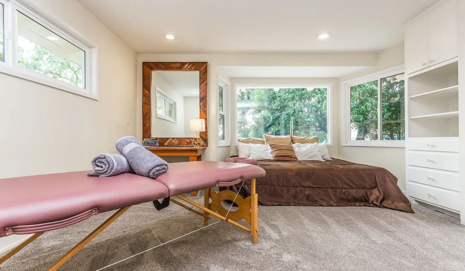 Dormitorio con una cama grande, una camilla de masaje rosa y ventanas con vista a la vegetación.