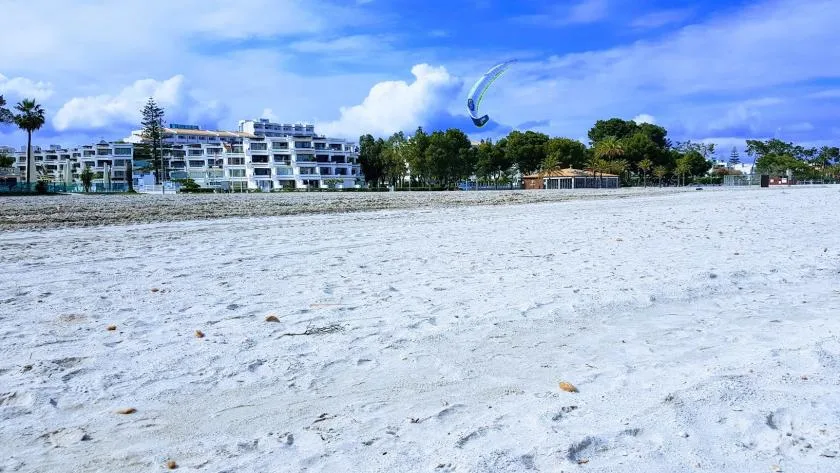 A sandy beach stretches toward waterfront buildings beneath a blue sky.