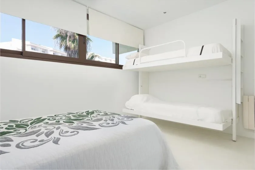 A bright bedroom with a patterned bed and a white bunk bed beside the window.