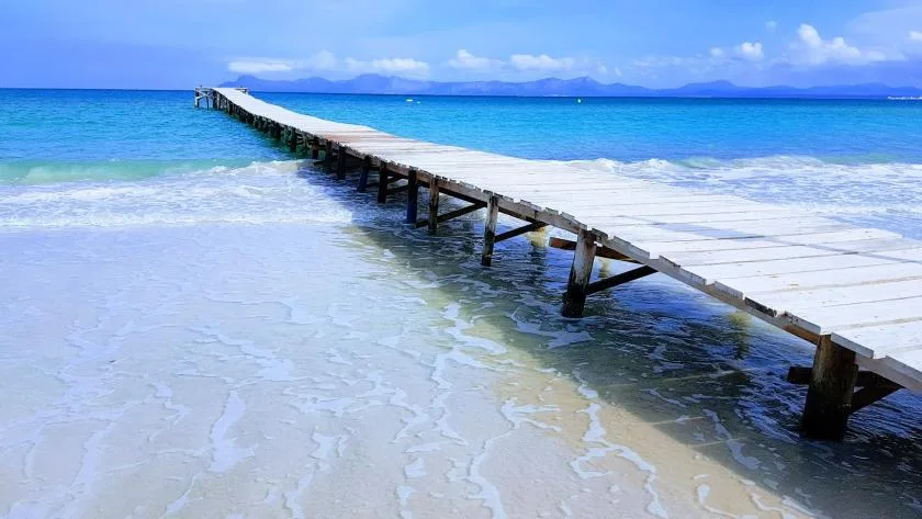 A wooden pier extends over clear turquoise water toward distant mountains.