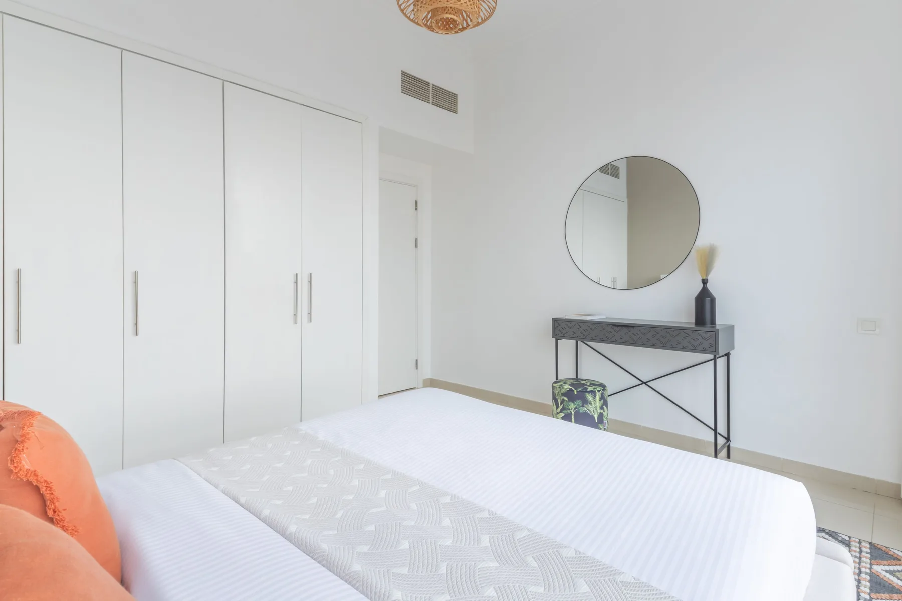 Un dormitorio luminoso con una cama, armarios blancos y un espejo redondo sobre una consola.