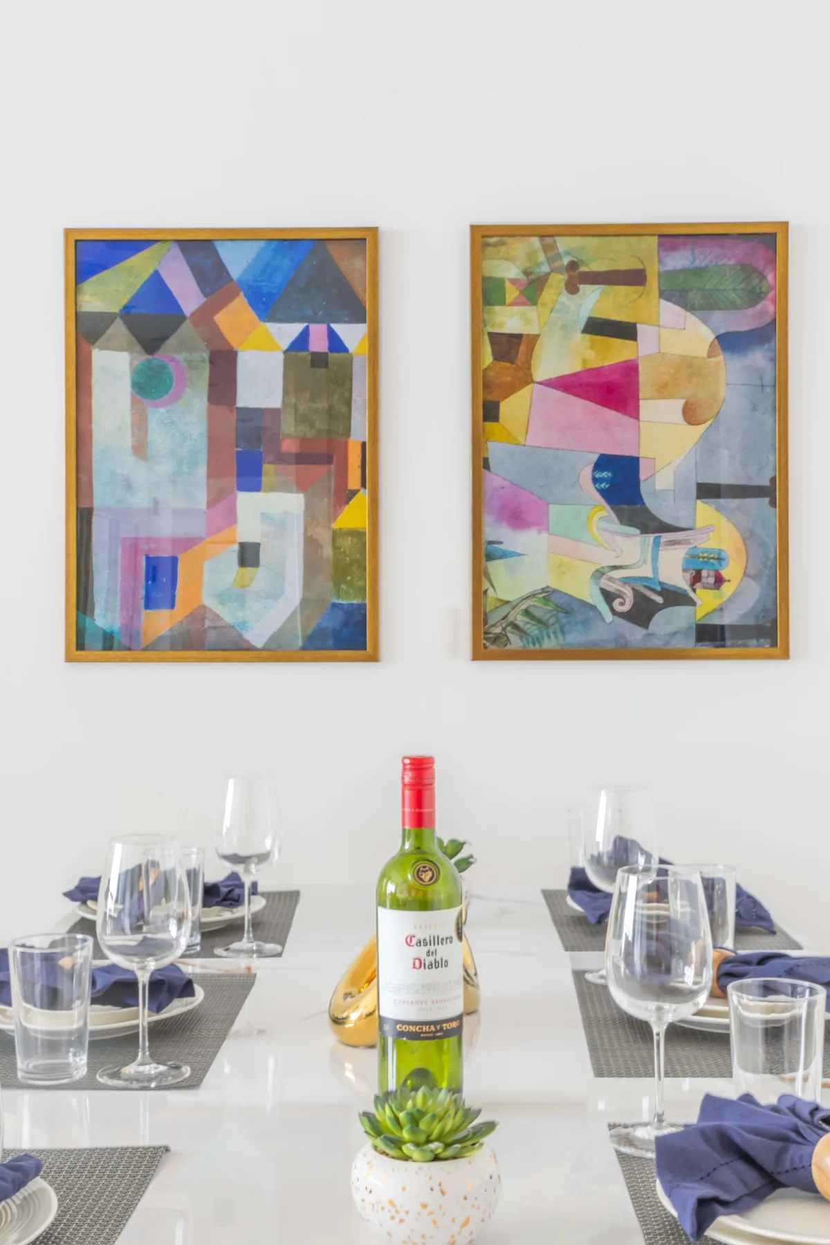 Una mesa de comedor preparada con vino y copas está bajo dos cuadros coloridos.