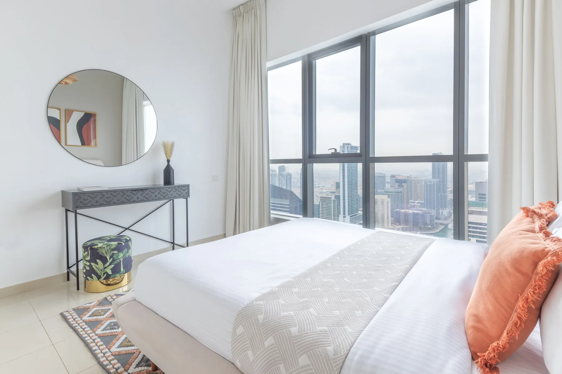 Un dormitorio con una cama bien arreglada junto a grandes ventanas con vistas a la ciudad.